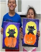 A young boy and girl stand side by side holding up their finished cat paintings.
