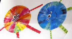 2 paper monsters are made out of tye-dye painted paper plates with frayed edges, multiple googly eyes, and folded strips of paper make up the arms and legs.