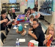 Children, some dressed in costumes, are gathered together around a table and enjoying bowls of chili and hotdogs.