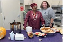 2 women stand together in the kitchen while preparing a pot of chili and some hot dogs. 