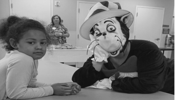 A girl leaning on a table with a person in a cat in the hat costume. A woman is in the background.